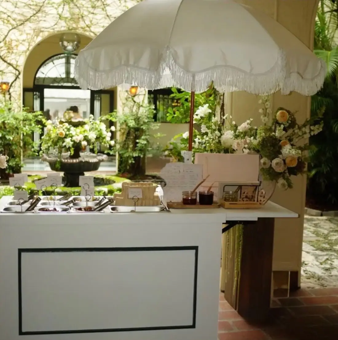 The Charcuterie Carte: a white cart with labelled stainless bins beneath a fringed white umbrella, in a courtyard with a fountain and flowers