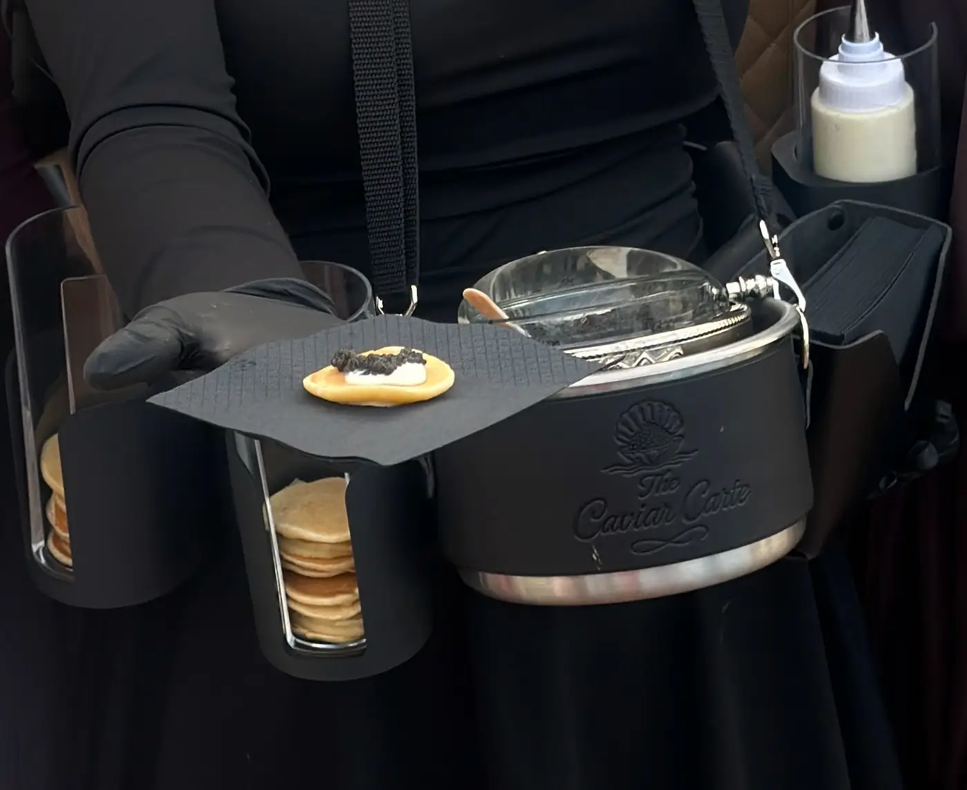 The signature service belt: a gloved hand offers a blini on a black napkin beside the embossed Caviar Carte tin, blini stacks and a bottle of crème fraîche