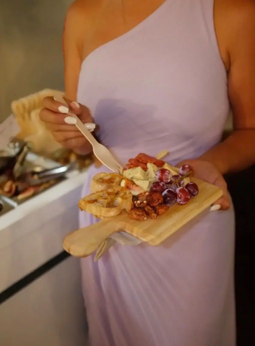 A guest in a lilac dress holds a finished board with grapes, cheeses and candied pecans